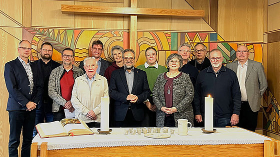 Das Gruppenbild zeigt dreizehn der sechzehn Personen des Kirchenvorstands der Evangelisch-methodistischen Kirche. Sie stehen an der Frontseite der Kapelle versetzt nebeneinander vor der bunt gestalteten Wand, an der ein Kreuz befestigt ist.