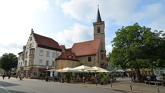 Im Vordergrund ein Teil eines Platzes mit Gastronomie und Bäumen. Im Hintergrund eine hauptsächlich aus Natursteinen gebaute Kirche mit Turm. Sie hat ein rotes Satteldach. Ein Fenster ist rechteckig. Die anderen Fenster haben Spitzbogen.