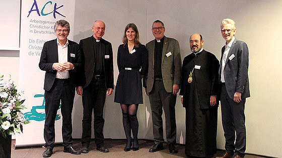 n einer Raumecke stehen fünf Männer und eine Frau. Der Katholik und der Orthodoxe sind in Amtstracht gekleidet. Der Anglikaner hat ein Kollarhemd an. Die anderen drei Personen sind nicht als Geistliche erkennbar.