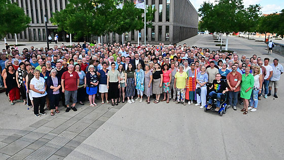 Gruppenfoto einer großen Anzahl von Menschen, die vor einem modernen Gebäude stehen. Die Personen sind unterschiedlich alt und tragen verschiedene Kleidung.