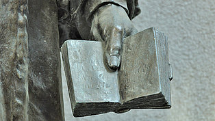 Nahaufnahme einer Bronzeskulptur, die eine Hand zeigt, die eine geöffnete Bibel hält. Die Skulptur ist detailliert und zeigt die Textur des Metalls und die Falten des Buches. Der Hintergrund ist unscharf und zeigt eine graue Wand.