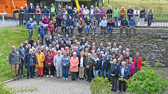 Das Bild zeigt die Konferenzmitglieder, die für ein Gruppenfoto posieren. Sie stehen im Freien vor einer niedrigen Steinmauer. Im Hintergrund ist ein gelber Reisebus zu sehen. Die Menschen sind unterschiedlich alt und tragen verschiedene Kleidung.