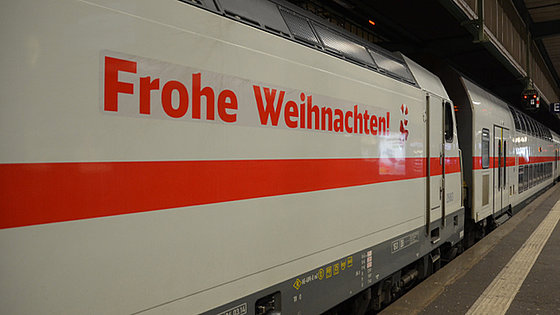 Das Bild zeigt eine Lokomotive der Deutschen Bahn im klassischen Weiß mit rotem Seitenstreifen. An der Seite trägt sie einen in Rot angebrachten Schriftzug. Darauf steht der sogar mit einem Ausrufezeichen versehene Gruß: Frohe Weihnachten!