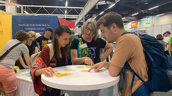 Im Vordergrund ein weißer Stehtisch mit drei Personen, die gelbe Quadrate mit Symbolen darauf so zusammenlegen versuchen, dass sich ein ganzes Bild ergibt. Weiter hinten links ein größerer Tisch mit dem Legespiel und weiteren drei Personen.