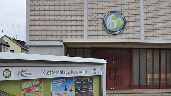 Zu sehen ist die Außenansicht der Kletterhalle. Ein Betonbau dessen Fassade zum Teil eine Backsteinoptik aufweist. Über den Eingang hängt das Logo der Kletterhalle. Dort steht „h 3” in weißer Schrift auf grünem Hintergrund.