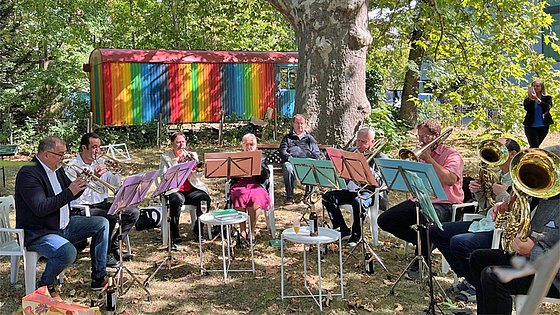 In einem Garten unter einem großen alten Laubbaum spielt ein achtköpfiger Posaunenchor: zwei Frauen und sechs Männer. Im Hintergrund steht ein bunt bemalter Wagen.