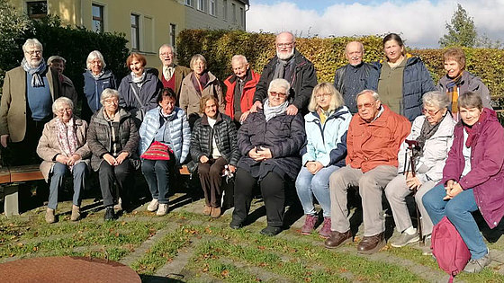 20 meist ältere Menschen im Freien, die in Jacken oder Mäntel gehüllt sind, sitzen auf einer Bank oder stehen dahinter. Die Mehrheit sind Frauen. Im Hintergrund stehen Hecken und eines der Beherbungshäuser. Die Sonne scheint.
