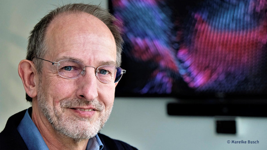 Klaus Ulrich Ruof mit kurzem Haar, hoher Stirn, randloser Brille und kurzem Vollbart schaut freundlich in die Kamera. Im Hintergrund ist leicht verschwommen ein Wandbildschirm zu erkennen.