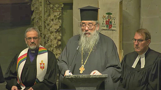 Im Vordergrund rechts ein in Dunkelgrau gekleideter Mann mit schwarzer Kopfbedeckung, langem Vollbart, Brustkreuz und Brille. Er steht vor einem Pult und spricht in ein Mikrofon. Im Hintergrund links steht ein Mann in schwarzem Talar und bunter Stola.
