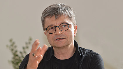 Porträt von Thomas Roscher mit kurzen, grau melierten Haaren und Brille. Er trägt ein schwarzes Hemd und gestikuliert mit der Hand. Der Hintergrund ist unscharf und hell.