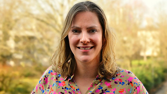 Eine Frau, Mittelscheitel, schulterlanges dunkelblondes Haar, bunte Bluse, schaut und lächelt in die Kamera. Im unscharfen Hintergrund ein Weg, Hecken und blattlose Bäume.