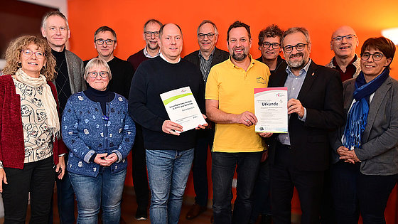 In zwei Reihen hintereinander stehen zwölf Personen, darunter drei Frauen, und sehen in die Kamera. Im Hintergrund eine orangefarbene Wand, die durch eine Stehlampe erhellt wird.