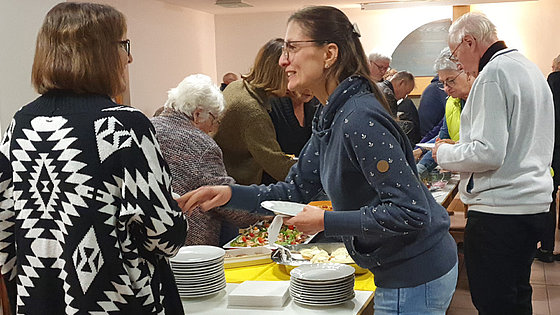 In einem Raum greifen Menschen auf ein Büfett zu. Im Vordergrund sind zwei Frauen mittleren Alters im Gespräch.