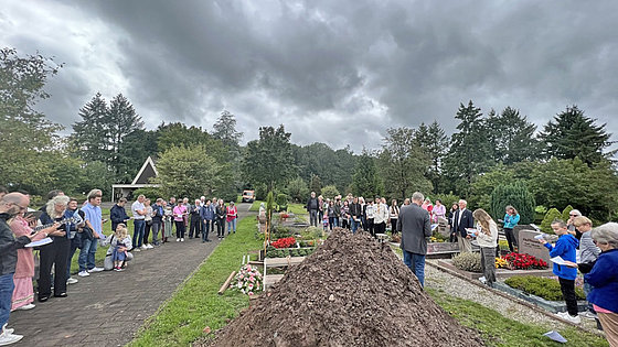 Auf einem Friedhof: Im Vordergrund ein Erdhaufen, dahinter das ausgehobene Grab und ein älterer Mann in Jackett und Jeans. Links auf einem Weg und rechts zwischen Gräbern etwa fünfzig Menschen unterschiedlichen Alters in meist farbiger Kleidung.