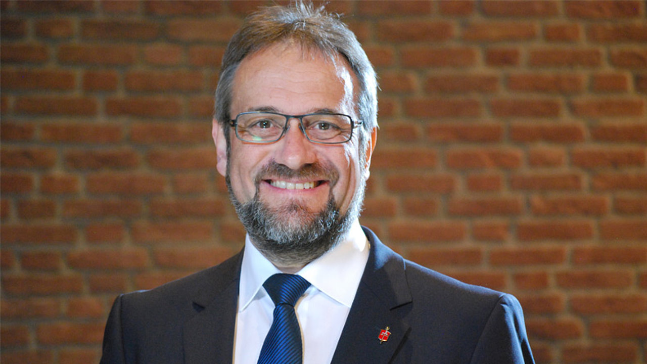 Portrait von Bischof in Ruhe Harald Rückert. Ein lächelnder Mann im fortgeschrittenen Alter mit Brille, Vollbart und dunklem Anzug. Er trägt eine blaue Krawatte und ein kleines Abzeichen am Revers. Der Hintergrund ist eine rote Backsteinmauer.