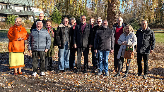 Unter herbstlich gefärbtem Laub einer Weide stehen 13 Personen in Winterkleidung. In der Mitte steht Bischof Werner Philipp. Alle blicken freundlich in die Kamera. Links im Hintergrund steht ein Gebäude.