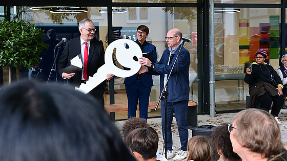 Christof Voigt, dunkler Anzug, weißes Hemd, rote Krawatte, nimmt einen hellen, mannshohen Schlüssel von Martin Bühler entgegen. Im Vordergrund einige Hinterköpfe von Zuschauern. Im Hintergrund die Glasfassade des neuen Foyers.
