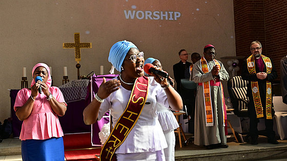Ghanaischer Festgottesdienst. Im Vordergrund links, vor den Stufen zum Altar, eine kleine Singgruppe mit drei ghanaischen Frauen in bewegter Pose. Rechts im Hintergrund stehen die Bischöfe Boafo aus Ghana und Rückert aus Deutschland mit farbiger Stola. 