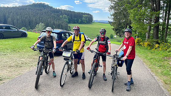 Vier Männer in Radlerkleidung mit Helm und Fahrrad schauen lächelnd in die Kamera. Sie stehen auf einer Straße, die hinter ihnen ins Tal führt. Grüne Wiesen und Wälder prägen die Landschaft. Der Himmel ist bewölkt. Nahe der Straße sind Autos geparkt.