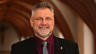 Portrait von Bischof Werner Philipp. Ein lächelnder Mannes mittleren Alters mit grau melierten Haaren und Bart. Er trägt einen dunklen Anzug und ein weißes Kollarhemd. Im Hintergrund sind unscharf Holzbögen zu sehen.