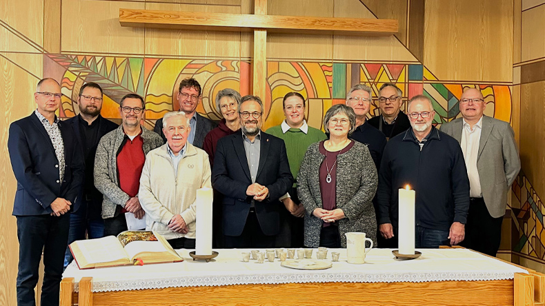 Das Gruppenbild zeigt dreizehn der sechzehn Personen des Kirchenvorstands der Evangelisch-methodistischen Kirche. Sie stehen an der Frontseite der Kapelle versetzt nebeneinander vor der bunt gestalteten Wand, an der ein Kreuz befestigt ist.