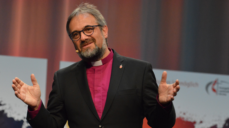 Bischof Harald Rückert, schwarzer Anzug, violettes Collar-Hemd, predigt einladend mit offener Handgeste auf einer Konferenzbühne. Den Hintergrund bildet ein verschwommen erscheinender, verschiedenfarbig lila, blau und grau angestrahlter Vorhang.