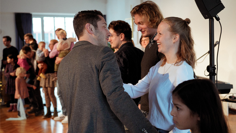 Eine Reihe von achtzehn Menschen von vorne rechts nach hinten links. Sie werden als Kirchenglieder aufgenommen. Vorne links ist Pastor Simon Kurfess, im Jackett, kurze Haare, Dreitagebart. Er begrüßt eine junge, fröhlich lachende Frau als neues Kirchenglied.