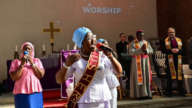 Ghanaischer Festgottesdienst. Im Vordergrund links, vor den Stufen zum Altar, eine kleine Singgruppe mit drei ghanaischen Frauen in bewegter Pose. Rechts im Hintergrund stehen die Bischöfe Boafo aus Ghana und Rückert aus Deutschland mit farbiger Stola. 