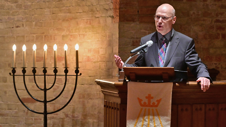 Links der siebenarmige jüdische Leuchter. Rechts steht Pastor Michael Putzke auf der Kanzel und spricht ins Mikrofon. Er hat eine Brille und trägt einen grauen Anzug mit bunter Krawatte. Er schaut ins Publikum und hebt dabei seine rechte Hand.