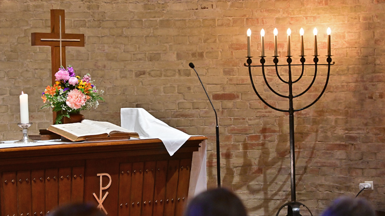 Links der Abendmahlstisch in dunklem Braun, darauf ein Kreuz und davor Blumen und Bibel. Links neben der Bibel eine weiße brennende Kerze. Rechts neben der Bibel Brot und Kelch zugedeckt. Rechts steht die Menora. Im Hintergrund eine Backsteinmauer.
