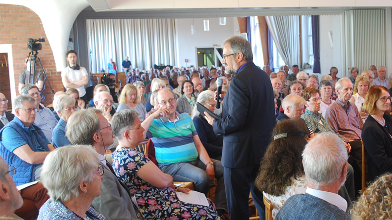 Ein Gottesdienstraum, in dem viele Menschen im Halbrund sitzen. Mittendrin steht Bischof Harald Rückert. Er hält ein Mikrofon in der Hand und blickt die ihm am nächsten sitzenden Personen an. Im Hintergrund eine Kamera zur Übertragung des Livestreams.