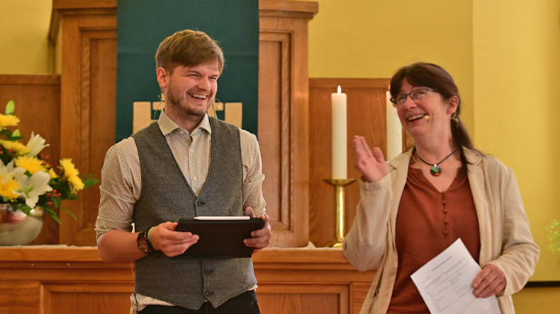 Im Vordergrund links ein junger bärtiger Mann, Melle, mit Tablet und rechts eine junge Frau, Iffland, mit Papier, beide einander anlächelnd. Im Hintergrund eine Kanzel und davor ein Abendmahlstisch mit Blumenschmuck und zwei brennenden Kerzen.