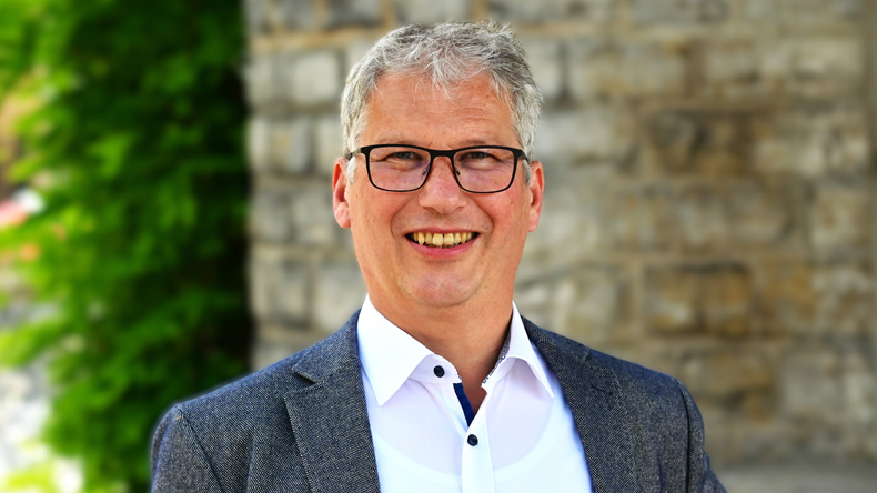 Matthias Kapp: volles graues Haar, Brille, ein Lächeln auf den Lippen, offenes helles Hemd und Jackett sieht direkt in die Kamera. Im Hintergrund eine Mauer aus Natursteinen und links eine Hecke.
