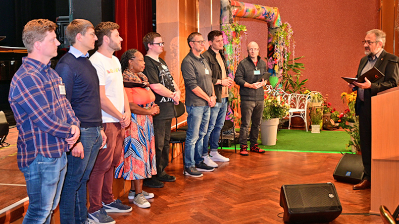 Links stehen sieben Männer und eine Frau in legerer Kleidung. Ihnen gegenüber steht der Bischof im Anzug. Im Hintergrund gibt es bunten Blumenschmuck.