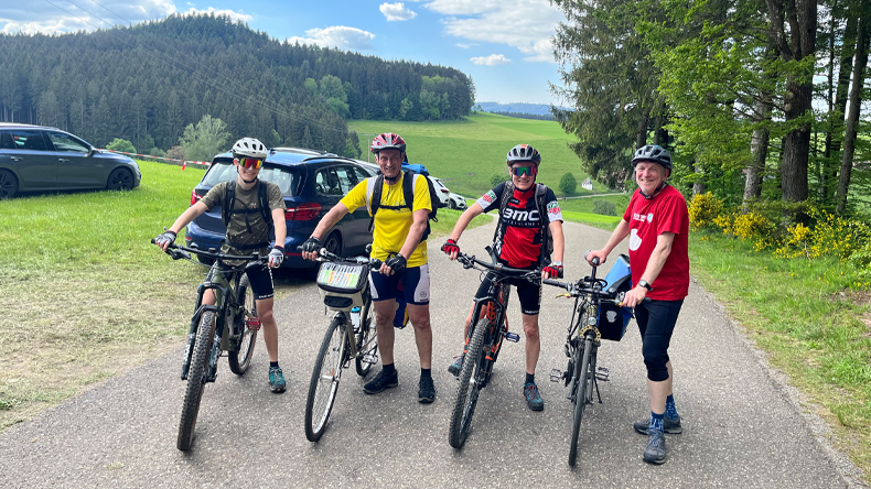 Vier Männer in Radlerkleidung mit Helm und Fahrrad schauen lächelnd in die Kamera. Sie stehen auf einer Straße, die hinter ihnen ins Tal führt. Grüne Wiesen und Wälder prägen die Landschaft. Der Himmel ist bewölkt. Nahe der Straße sind Autos geparkt.