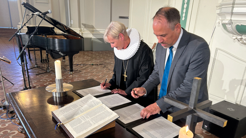 Fehrs: schwarzer Talar, bischöfliches Brustkreuz, weiße Halskrause. Brenner: grauer Anzug, weißes Hemd, blaue Krawatte. Beide sind dabei, die Erklärung auf dem Abendmahlstisch zu unterschreiben. Vor ihnen eine aufgeschlagene Bibel und zwei Kerzen.