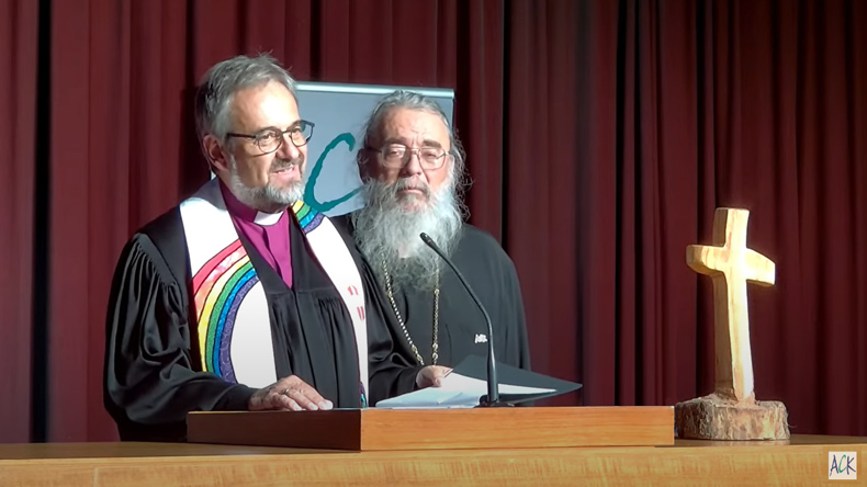 Harald Rückert – kurzer Vollbart, schwarzer Talar mit regenbogenfarbiger Stola und Kollarhemd – spricht die Gemeinde an. Radu Constatin Miron – langer Vollbart, dunkelgraues Gewand – hört ihm aufmerksam zu. Im Hintergrund ein purpurfarbener Vorhang.