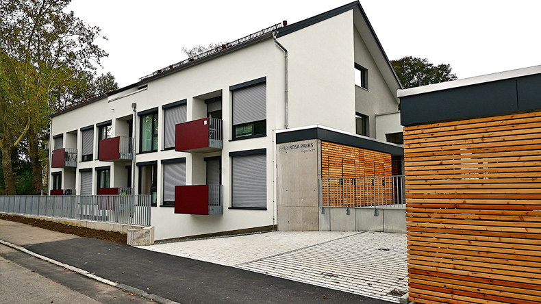 Eine breite Seite mit Fenstern und kleinen Balkonen sowie eine Giebelseite des neuen Wohnheims. Es ist ein zweistöckiges Gebäude plus Dachgeschoss. Direkt vor dem Wohnheim die gepflasterte Abfahrt zur Tiefgarage. Im Vordergrund die Hagstraße.