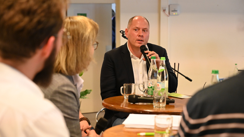 Stefan Weiland, Stirnglatze, kurze Haare, helles Hemd, dunkles Sakko, spricht in ein Mikrofon. Vor ihm sitzen Personen, die ihn ansehen. Auf dem Foto sind Rücken und Hinterköpfe abgebildet. Zwischen Weiland und den Personen stehen zwei runde Tische.