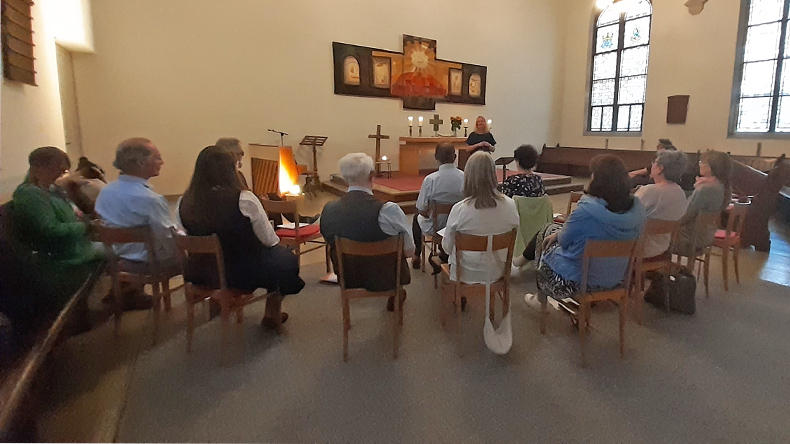 Rund ein Dutzend meist ältere Menschen sitzen auf Stühlen in einem Kirchensaal. Ihre Blicke sind auf eine Frau gerichtet, die sich im erhöhten vorderen Teil des Saals befindet. Auf dem Abendmahlstisch brennen Kerzen. Sonnenlicht erhellt den Raum.