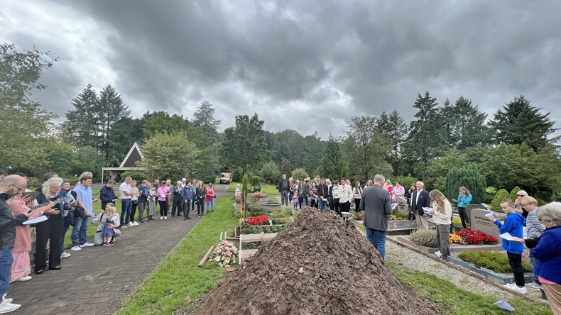 Auf einem Friedhof: Im Vordergrund ein Erdhaufen, dahinter das ausgehobene Grab und ein älterer Mann in Jackett und Jeans. Links auf einem Weg und rechts zwischen Gräbern etwa fünfzig Menschen unterschiedlichen Alters in meist farbiger Kleidung.