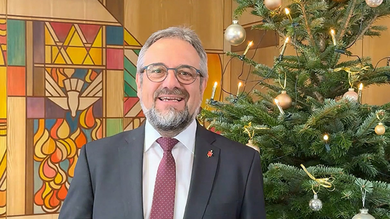 Ein Mann im schwarzen Jackett mit weißem Hemd und roter Krawatte. Rechts hinter ihm ist ein geschmückter Weihnachtsbaum. Links hinter ihm ist die bunt gestaltete Stirnwand eines Gottesdienstraums zu sehen.
