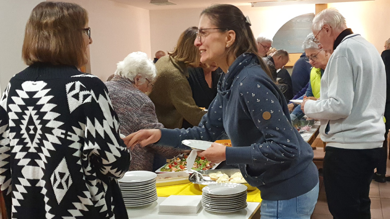 In einem Raum greifen Menschen auf ein Büfett zu. Im Vordergrund sind zwei Frauen mittleren Alters im Gespräch.