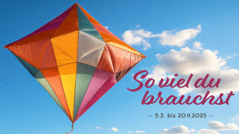 Vor dem leicht bewölkten Himmel hebt sich ein naher bunter Flugkörper aus Stoff ab. Er ist geometrisch zusammengesetzt aus zwei Pyramiden, die sich die Grundfläche teilen. Die untere Pyramide hat ihre Spitze unten. Text auf dem Bild: »So viel du brauchst«.