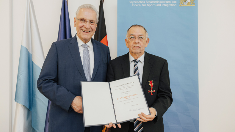 Zwei Herren halten zusammen die Verleihungsurkunde und blicken freundlich in die Kamera: links der Minister, rechts der Geehrte mit dem empfangenen Kreuz an der Brusttasche des Jacketts.