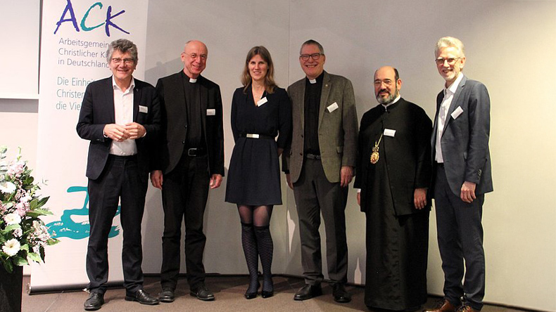 n einer Raumecke stehen fünf Männer und eine Frau. Der Katholik und der Orthodoxe sind in Amtstracht gekleidet. Der Anglikaner hat ein Kollarhemd an. Die anderen drei Personen sind nicht als Geistliche erkennbar.
