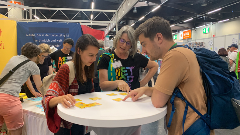 Im Vordergrund ein weißer Stehtisch mit drei Personen, die gelbe Quadrate mit Symbolen darauf so zusammenlegen versuchen, dass sich ein ganzes Bild ergibt. Weiter hinten links ein größerer Tisch mit dem Legespiel und weiteren drei Personen.