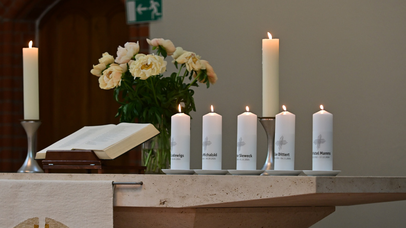 Auf einem Abendmahlstisch aus Stein befinden sich eine aufgeschlagene Bibel, ein Blumenstrauß, zwei hohe Kerzen und fünf mit Namensaufschrift versehene niedrigere Kerzen. Alle Kerzen sind angezündet.