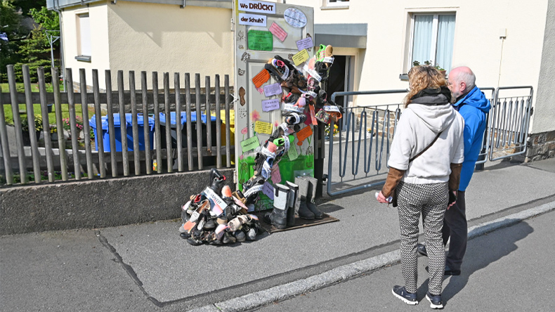 Zwei Personen stehen auf einer Straße und betrachten eine alte Tür, die an einem Holzzaun befestigt ist. Vor der Tür ist schräg ein Kreuz aus Schuhen festgemacht. Auf der Tür und auf dem Kreuz kleben Zettel mit Botschaften zum Nachdenken.