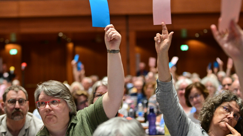 In einem großen lichtdurchfluteten Raum sitzen viele Menschen. Einige von ihnen halten blaue oder rosafarbene Stimmkarten in die Höhe.